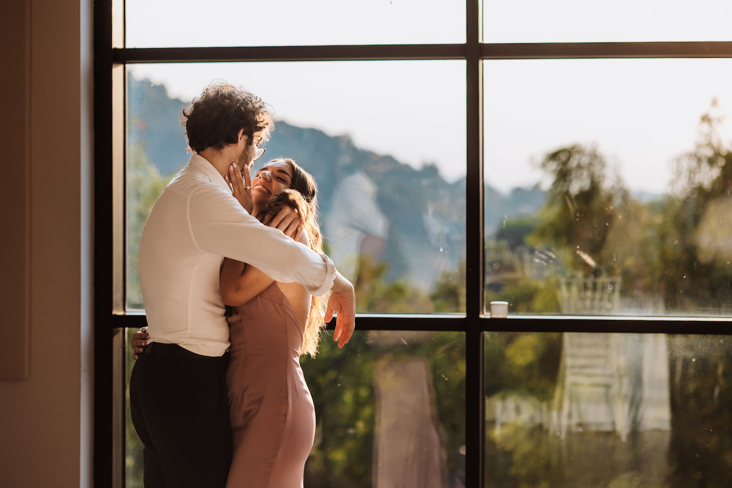 Couple embracing by a large window with a scenic mountain view in warm golden light.