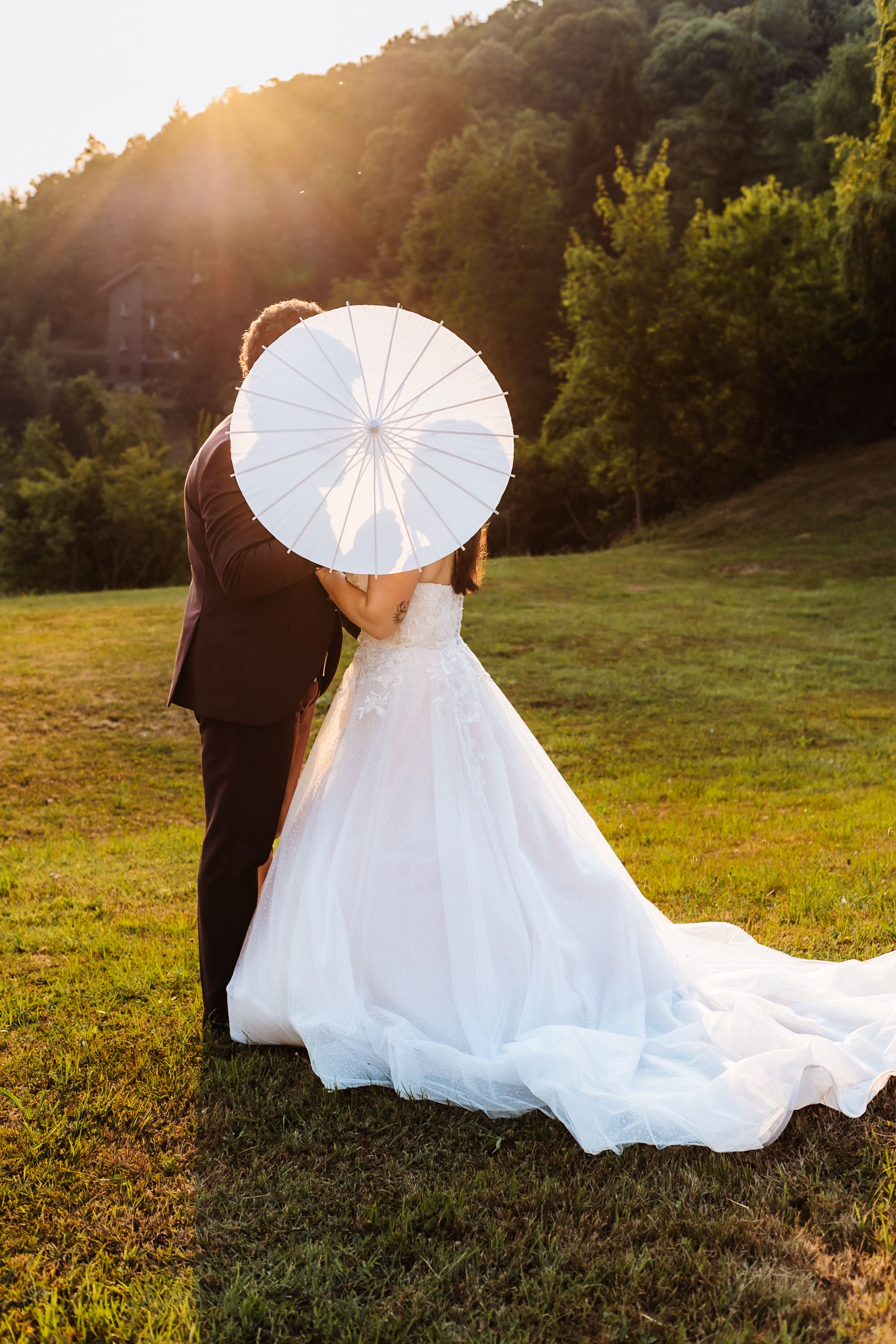 Bride and groom hiding behind a white parasol umbrella in a sunlit green field.