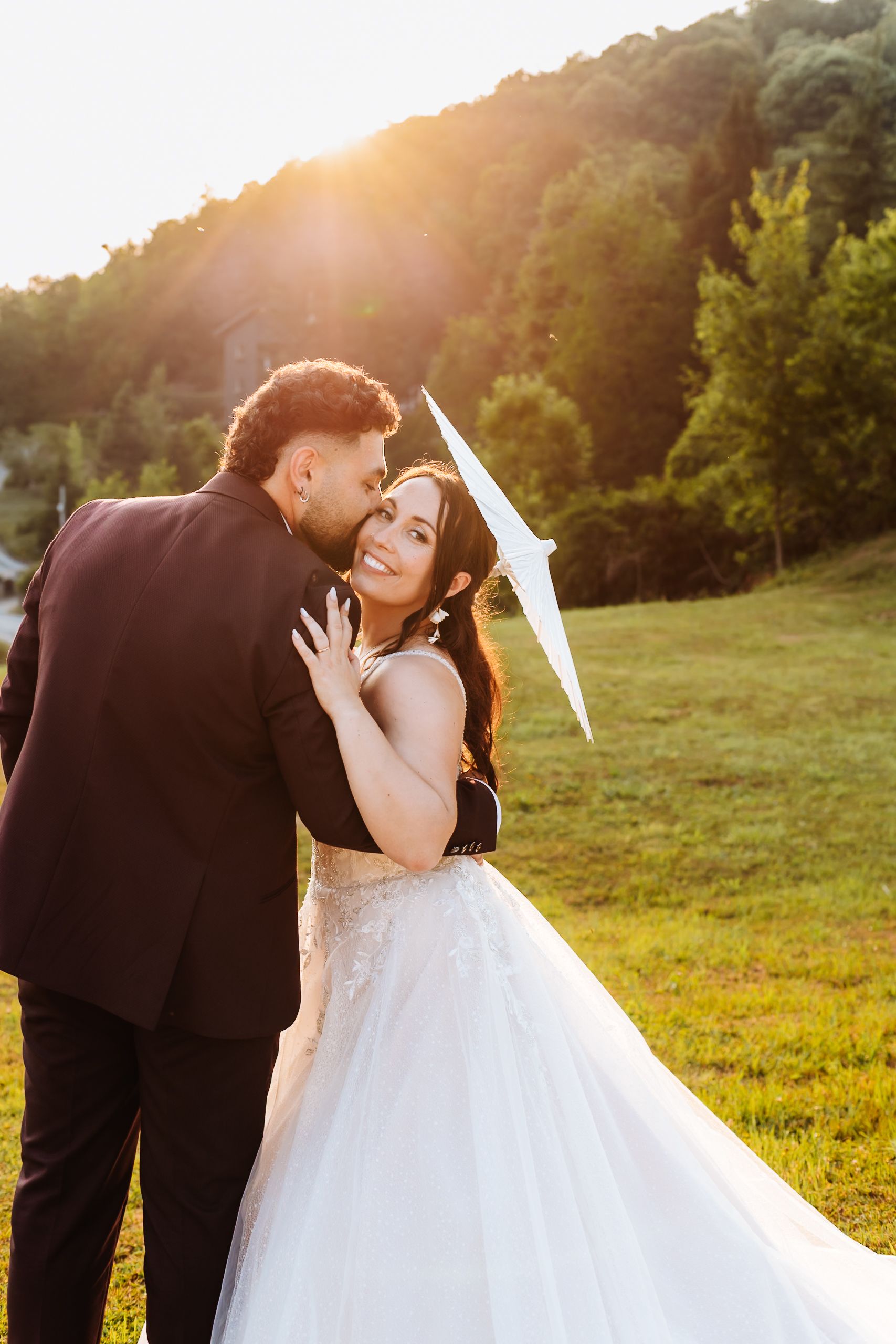 Bride in white gown smiles as groom embraces her during golden hour outdoor wedding portrait.