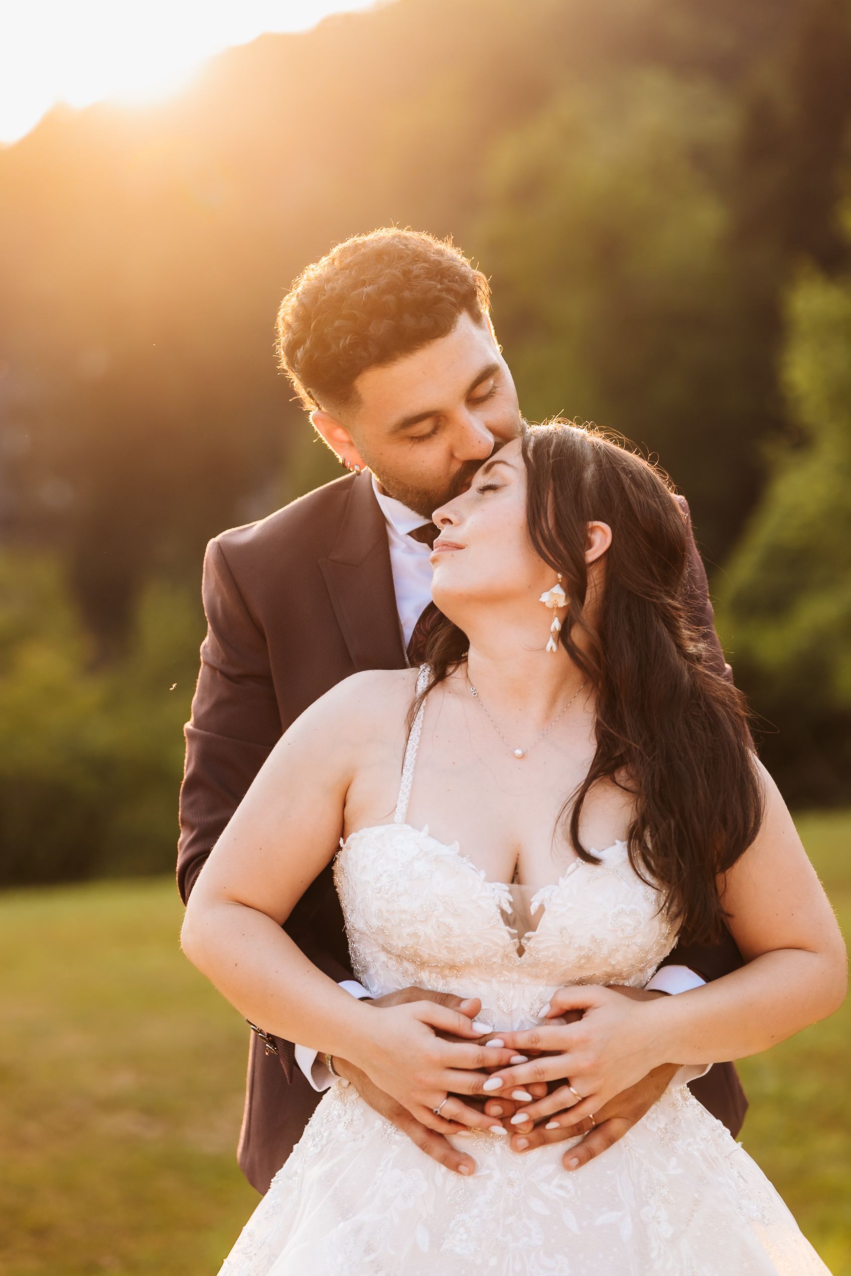 Bride and groom embrace at golden hour sunset, groom kissing bride's cheek in romantic outdoor wedding portrait.