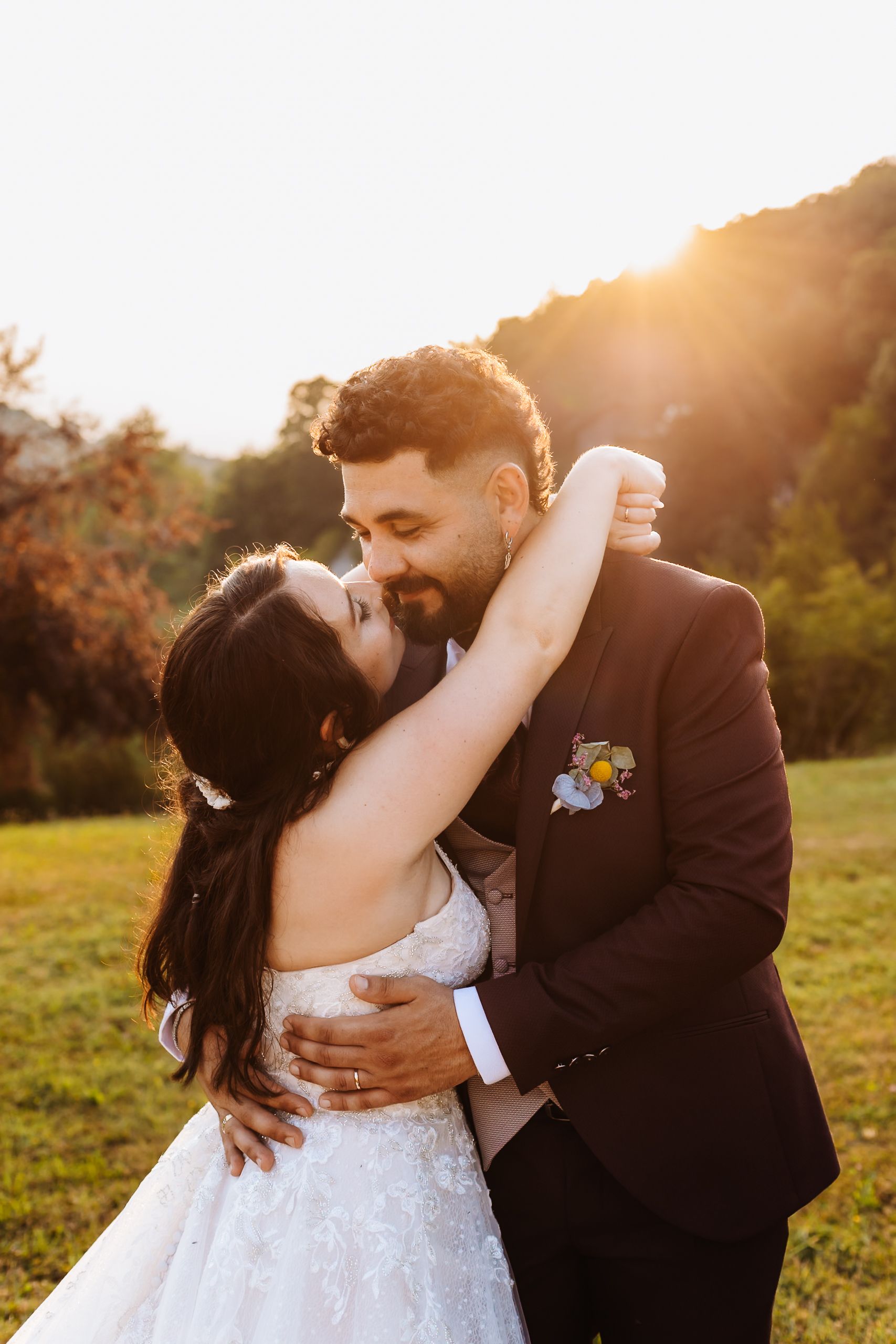 Bride and groom share a romantic embrace at golden hour with sunlight glowing behind them in a lush green field.
