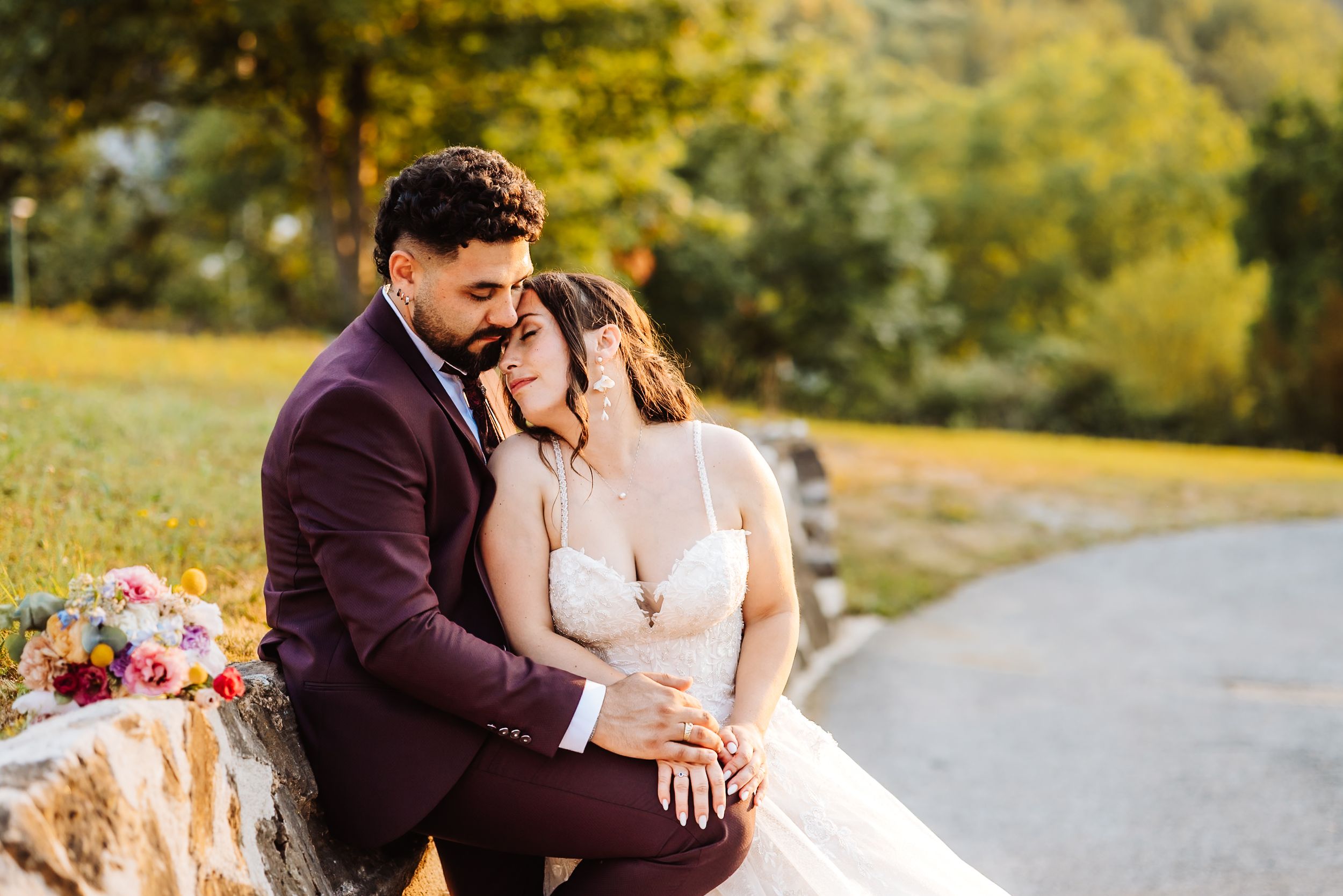 A couple in wedding attire shares a tender embrace outdoors, the groom in burgundy suit and bride in white gown.