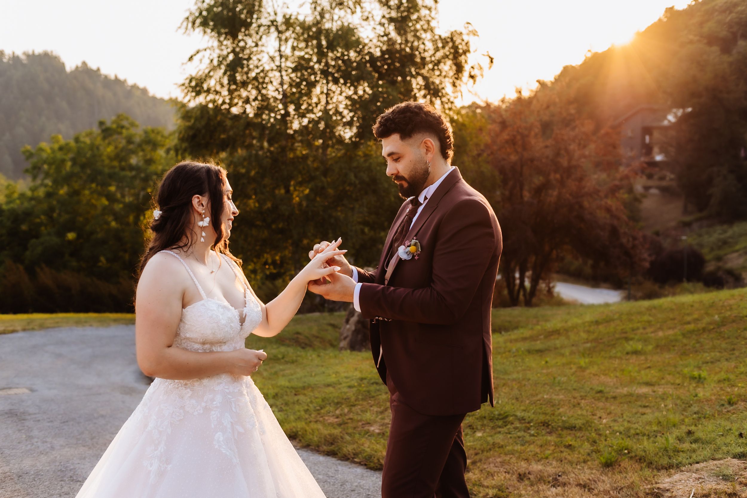 Bride and groom exchange rings at golden hour outdoors, surrounded by lush greenery and warm sunset light.