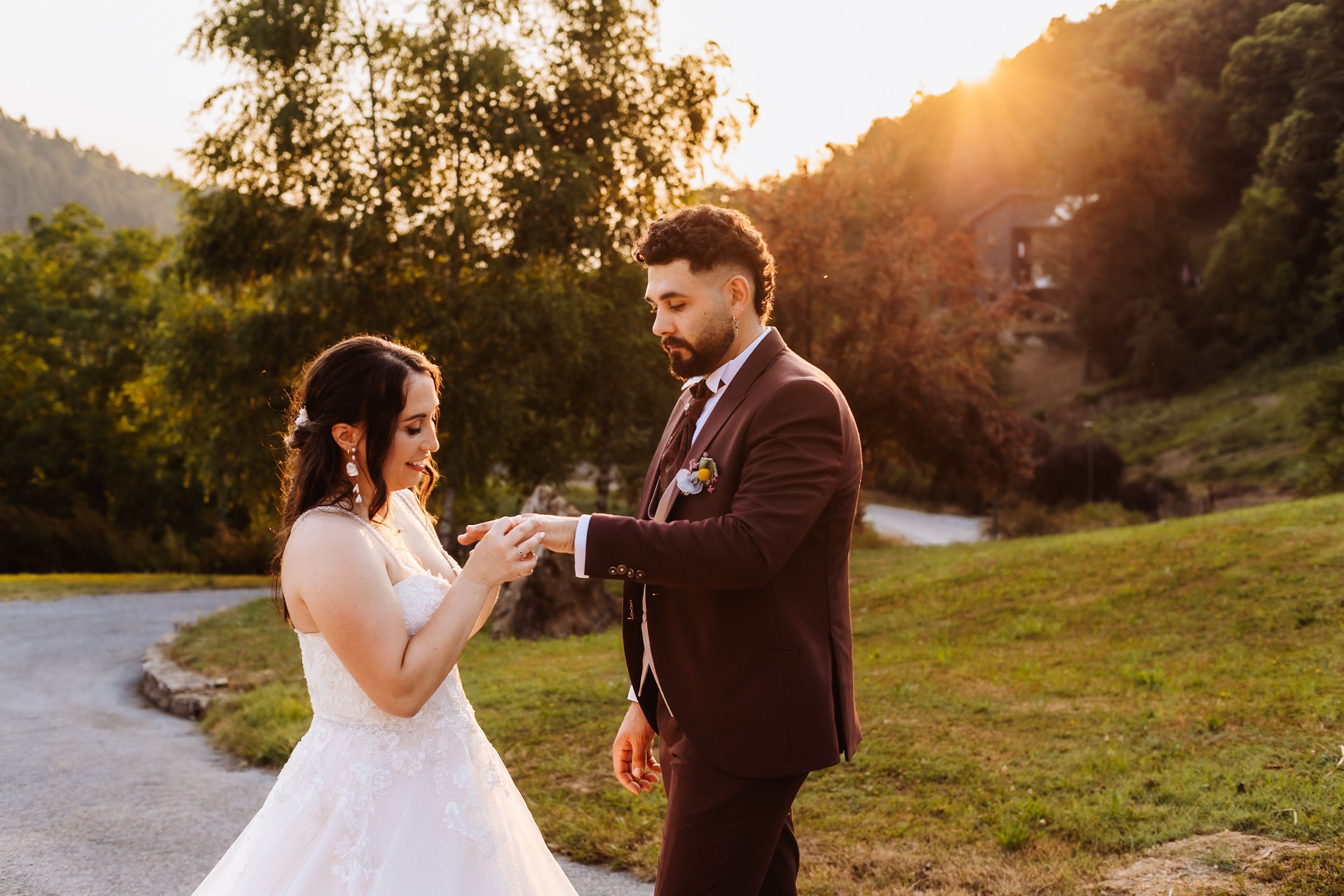 Groom placing a ring on bride's hand outdoors at golden sunset with green hills behind.