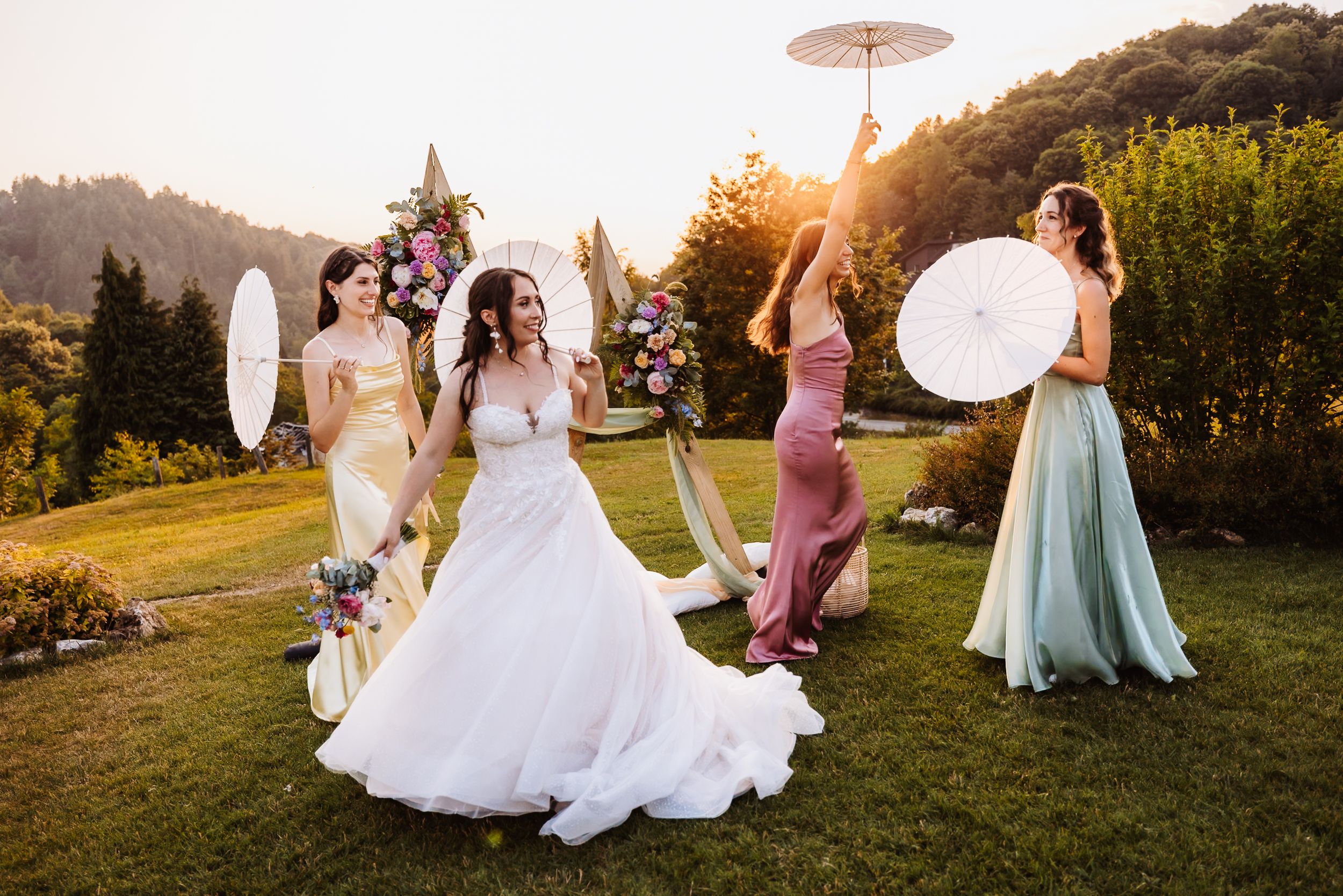 Bride in white gown poses with bridesmaids holding parasols at a sunlit outdoor wedding ceremony.