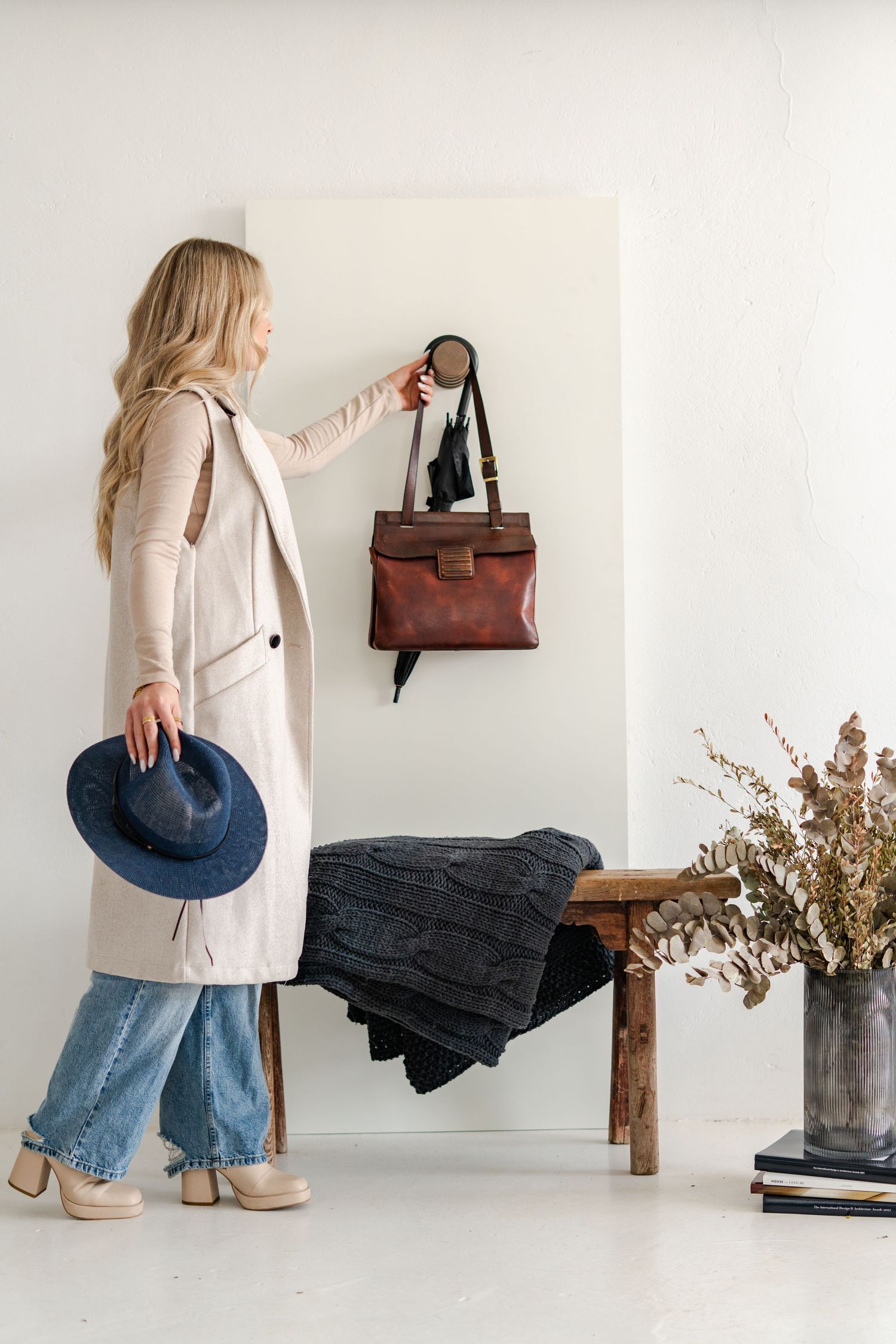 Blonde woman in beige coat and jeans hanging brown leather bag on wall hook in minimalist entryway.