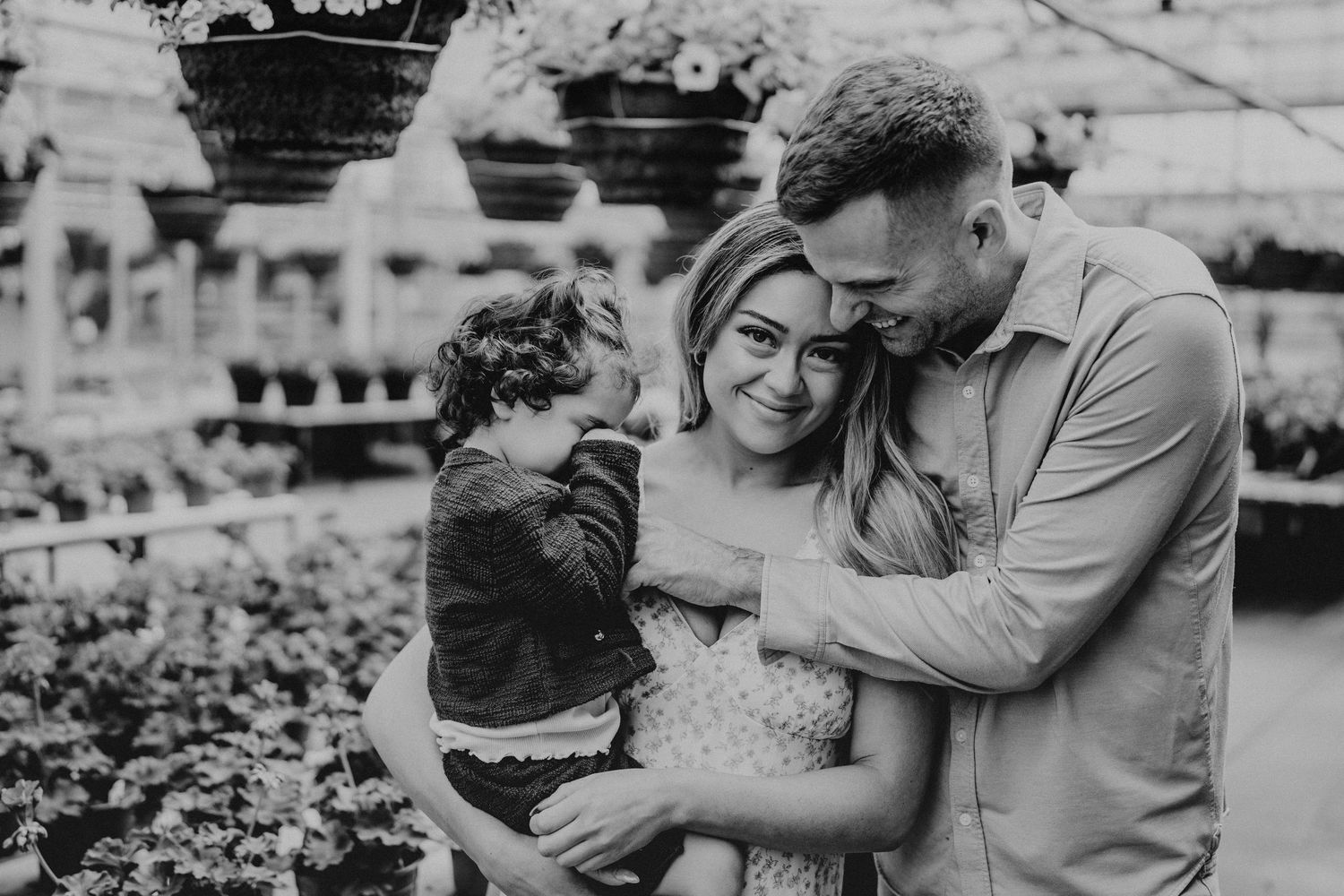 Black and white photo of a happy family with a toddler embracing in a lush greenhouse setting.