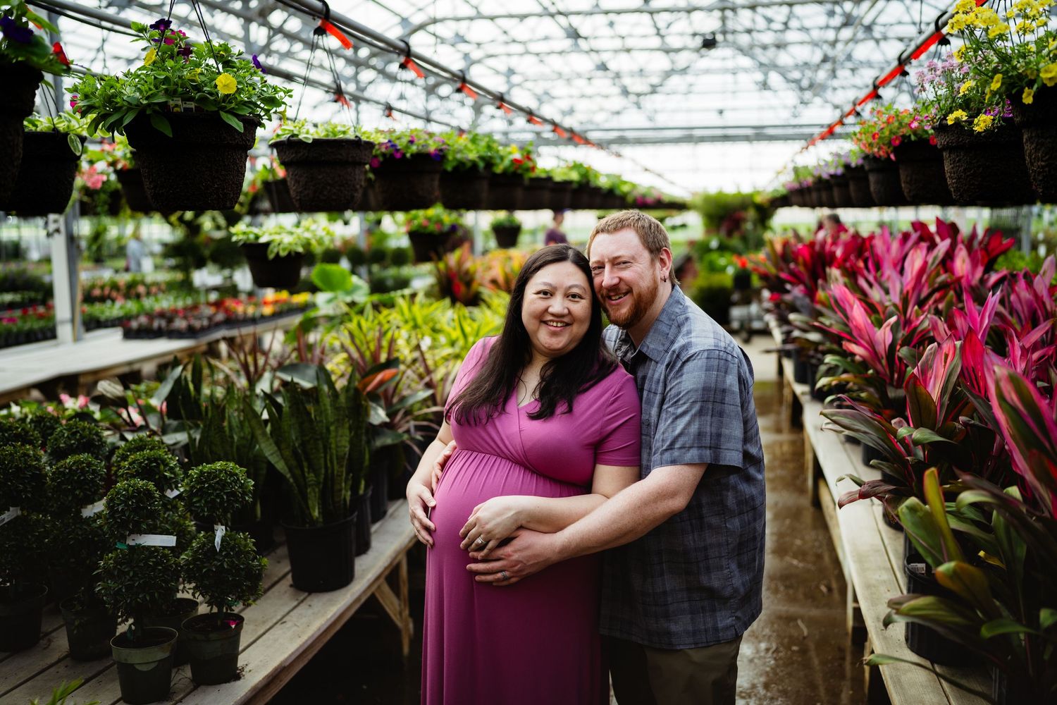 Pregnant couple smiling and embracing in a lush greenhouse surrounded by colorful tropical plants and hanging baskets.