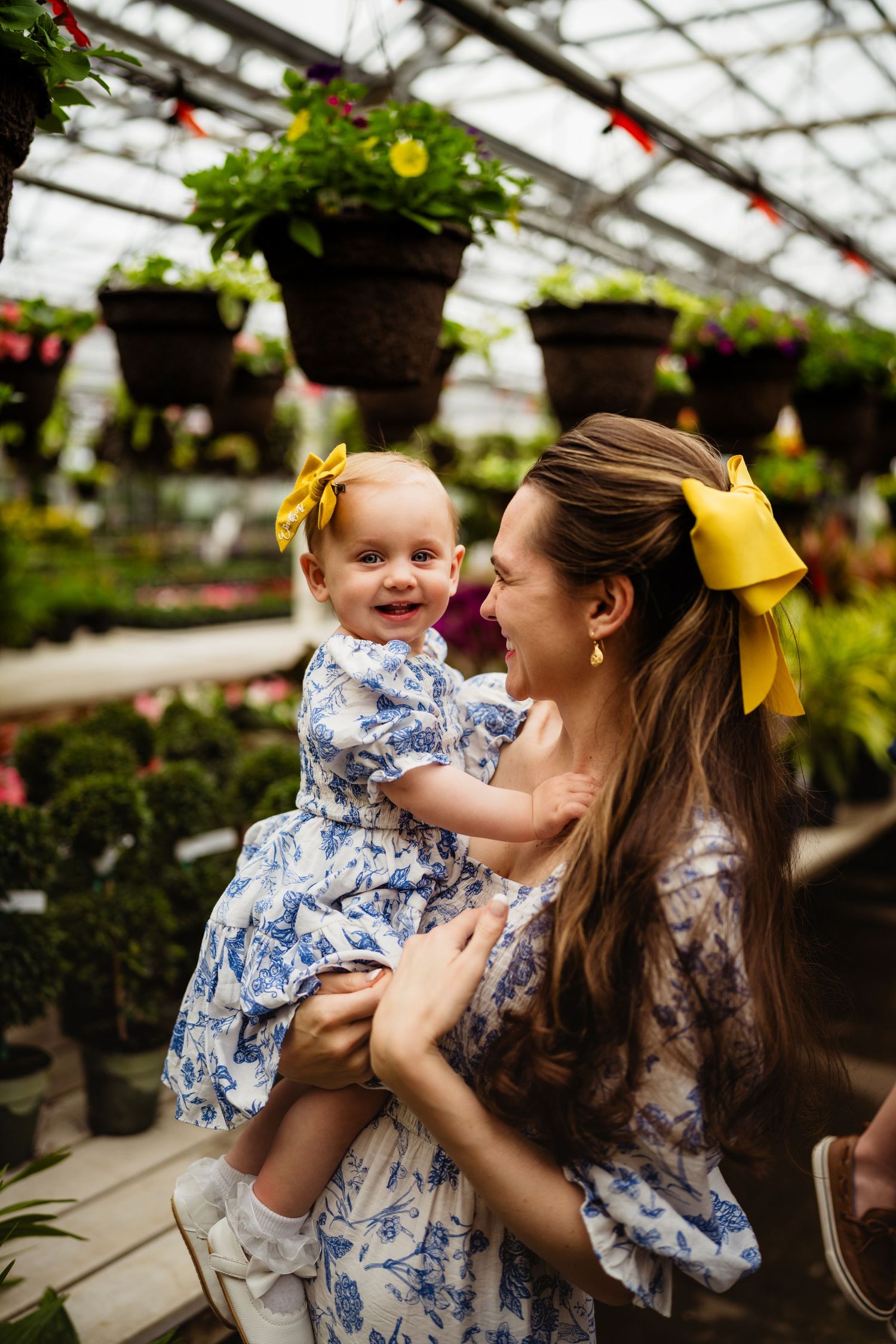 Mother and baby girl in matching blue floral dresses with yellow bows pose in a lush greenhouse.