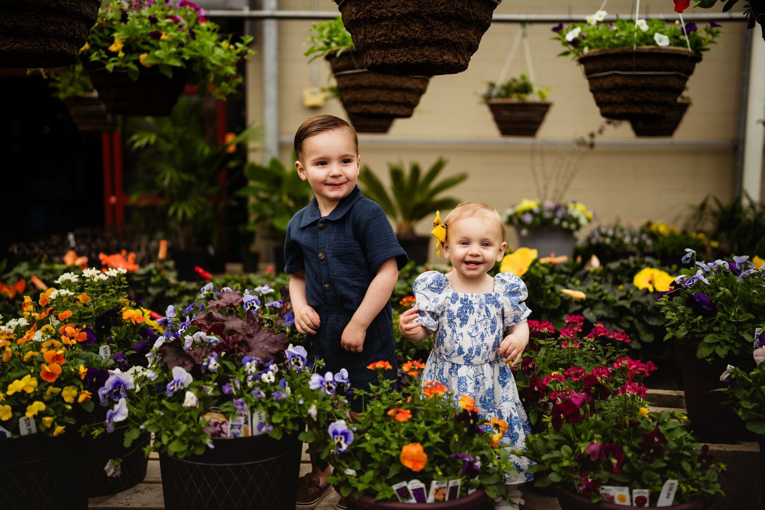 Two young children smile among colorful potted flowers and hanging baskets in a bright garden center.