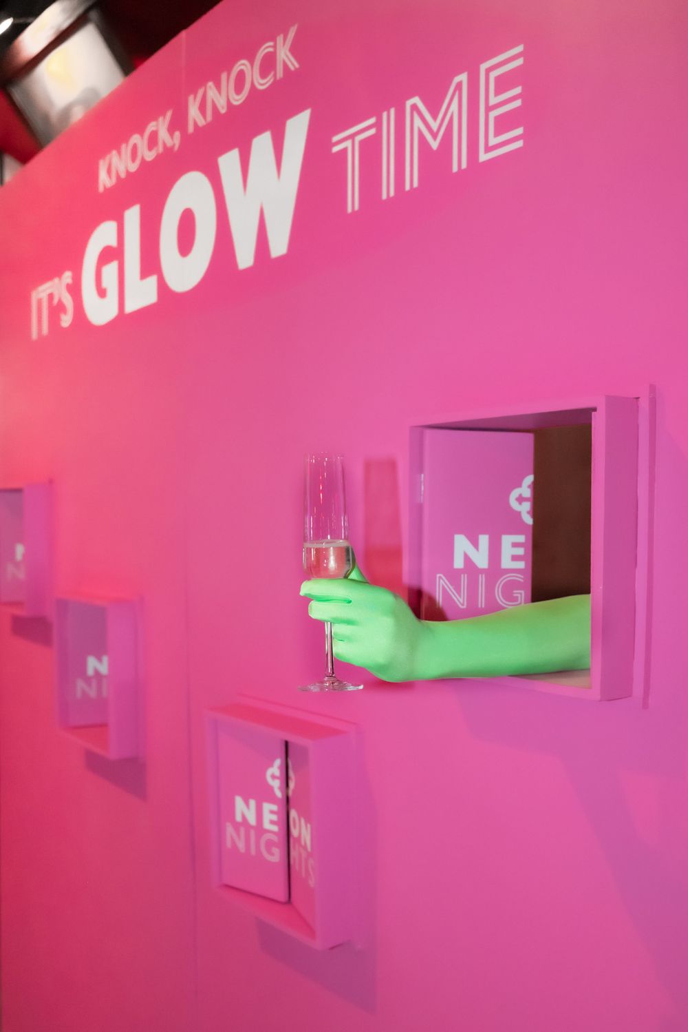 Pink wall display with neon sign reading 'It's Glow Time' and illuminated window cutouts.