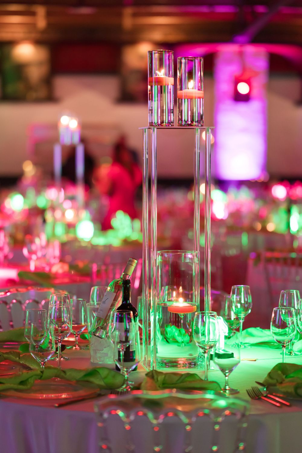 Tall glass centerpiece with glowing candles on an elegant reception table with dramatic pink and green lighting.