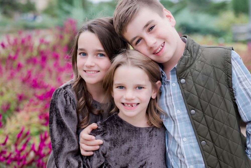 Three siblings pose together against a background of pink flowers in an outdoor portrait session.