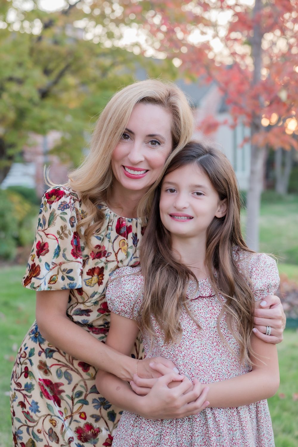 Parent and child wearing floral dresses embrace outdoors with fall foliage in background.