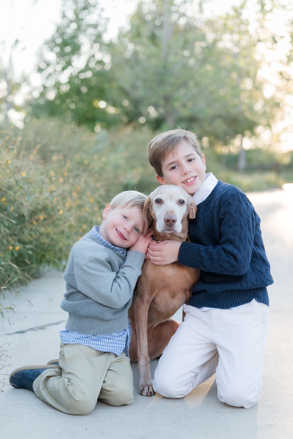Two children kneel beside their brown Weimaraner dog in outdoor family portrait.