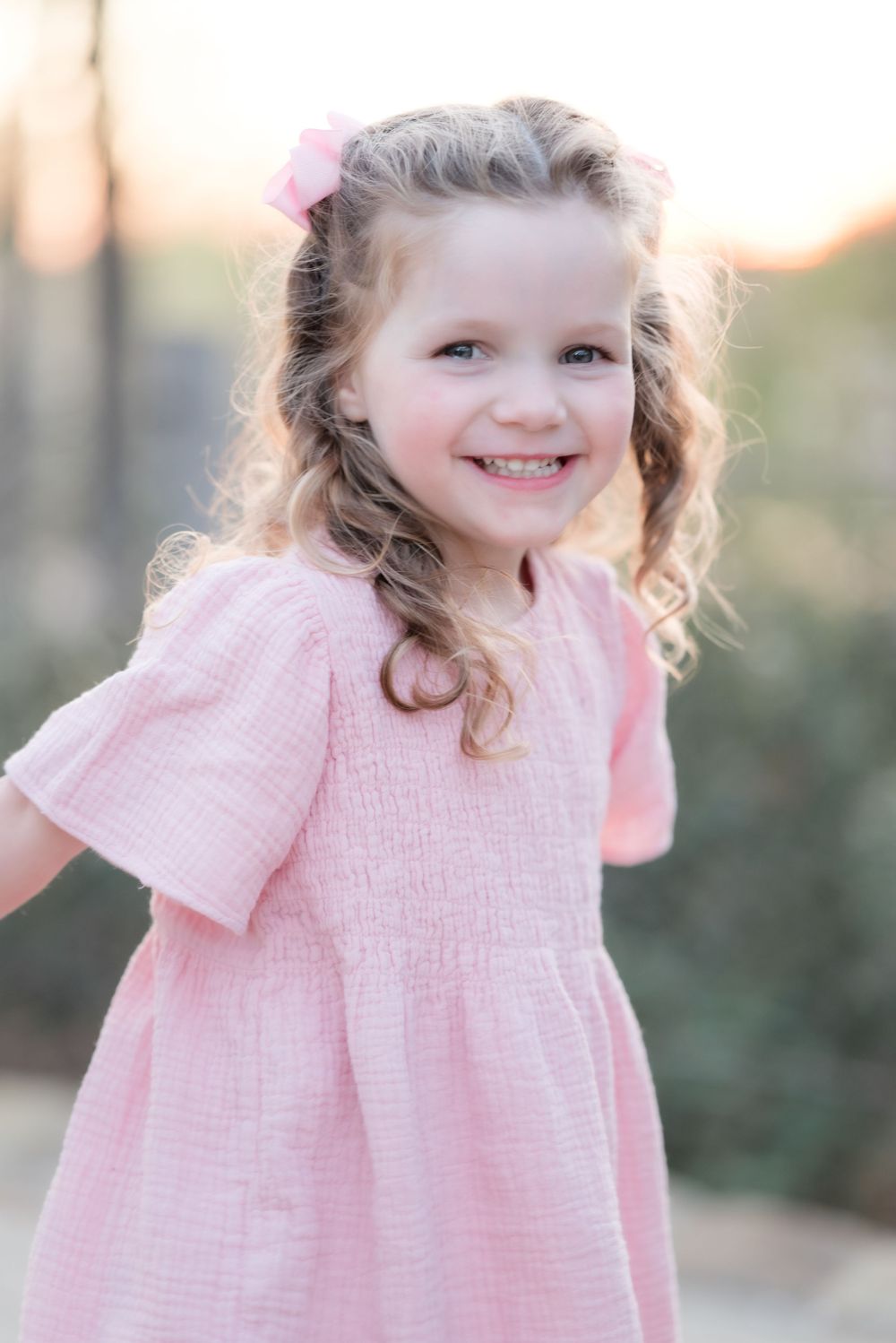 Portrait of a young child in a pink dress with curled hair and natural lighting outdoors.