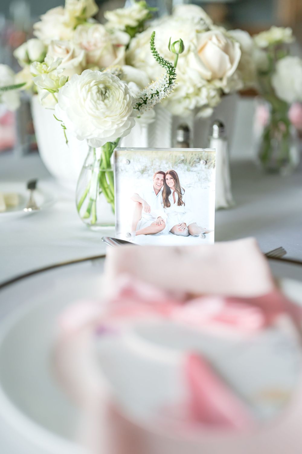 White floral centerpiece with roses in vase next to framed photo on elegant table setting.