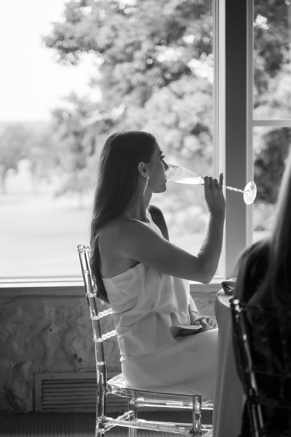Black and white photo of bride sipping champagne by window.