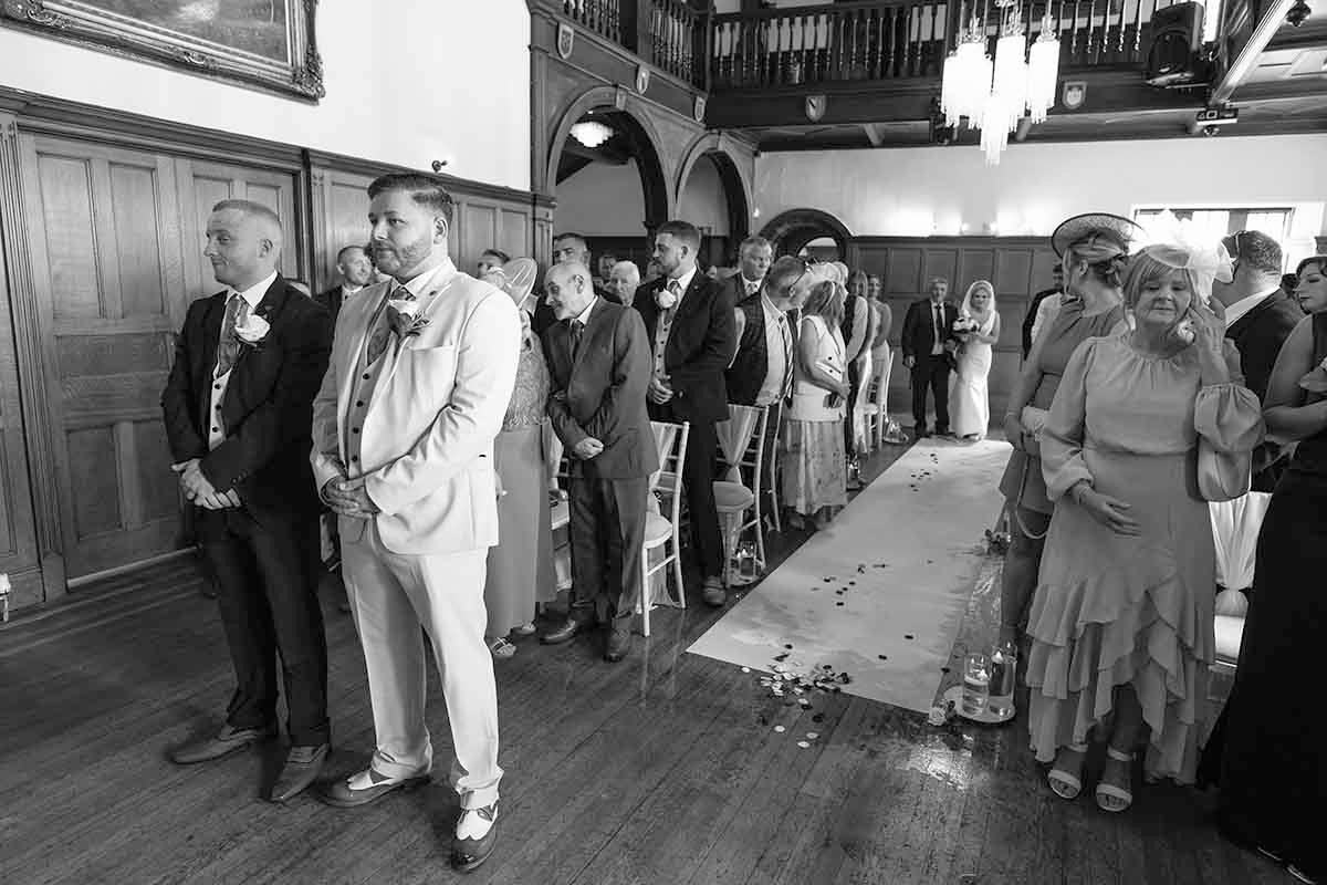 Black and white wedding ceremony photo showing guests standing in an elegant hall as the bride walks down the aisle.