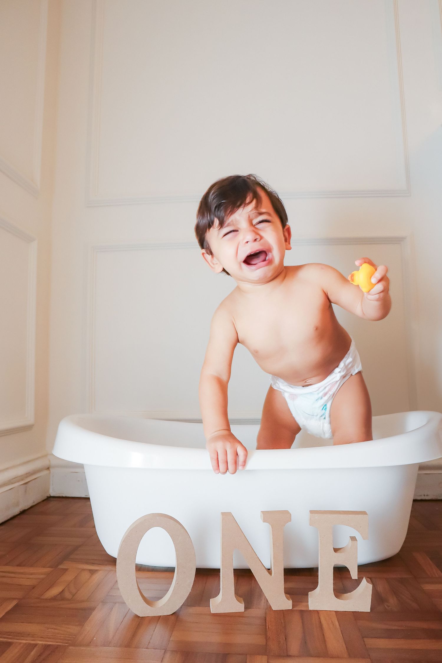Crying one-year-old baby in diaper standing in white bathtub with rubber duck and wooden ONE letters on hardwood floor.