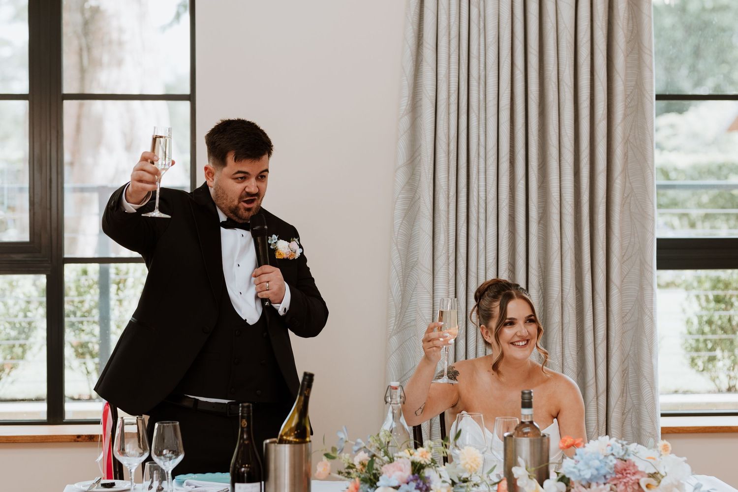 Groom in tuxedo raises champagne glass while bride smiles during wedding reception toast at elegant venue.