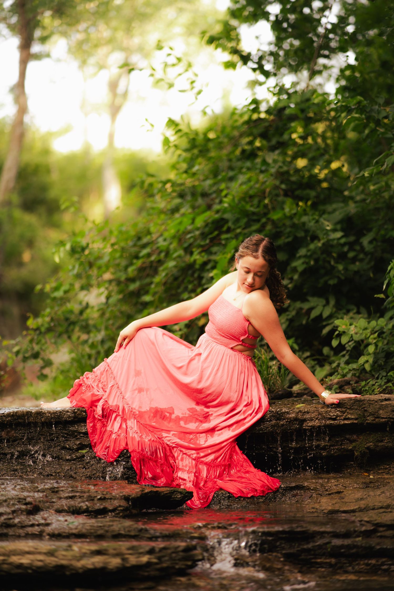 Young woman in flowing coral pink dress sitting gracefully on rocks beside a gentle stream in lush green forest.