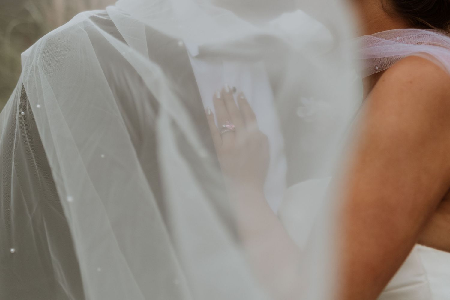A bride's veil draped over a groom's white shirt, with her hand gently resting on his back.
