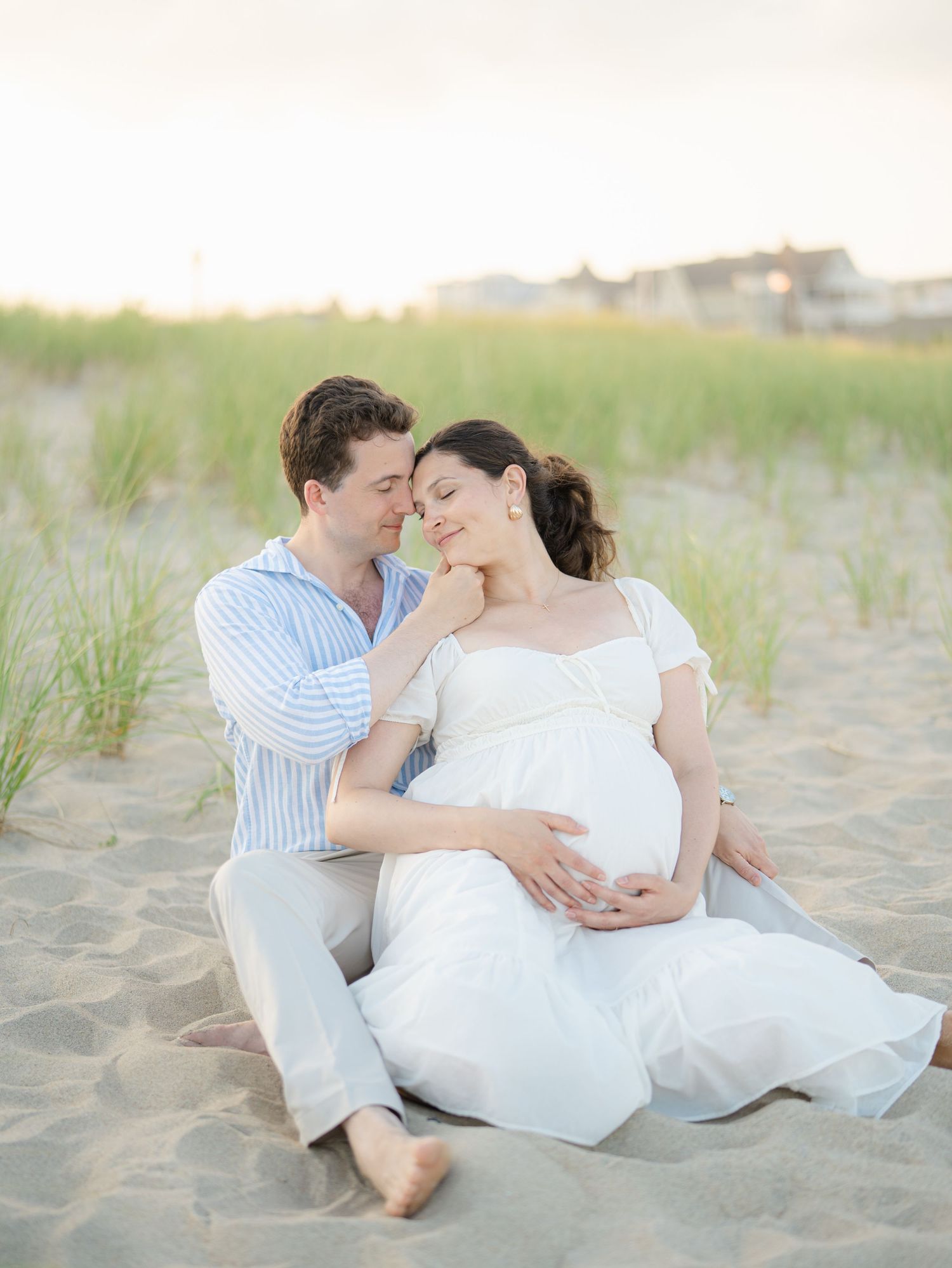 Husband snuggles pregnant wife while sitting on Ocean Grove beach at sunset Monmouth County New Jersey.
