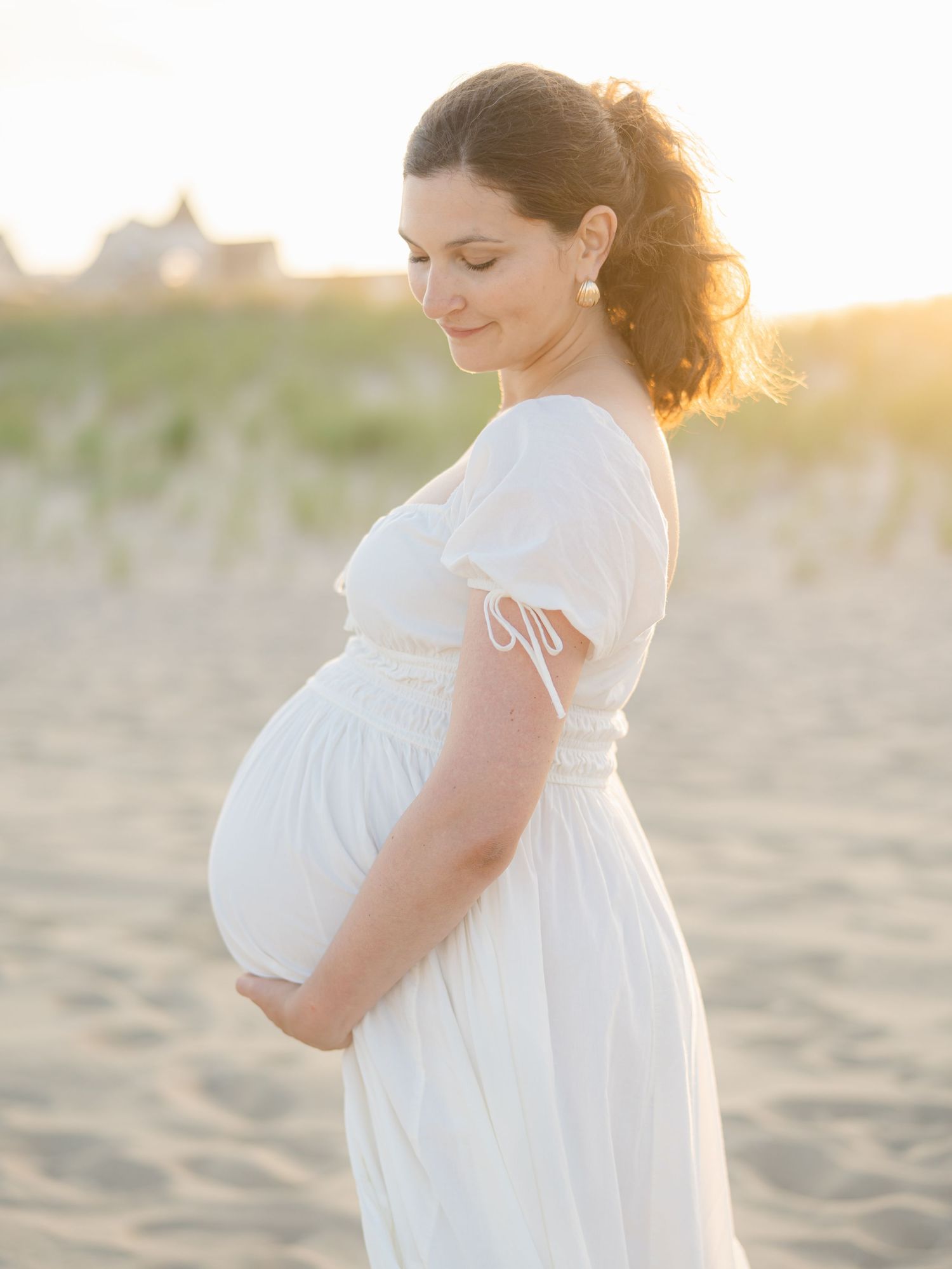 Pregnant mom in white flowing maxi dress standing in Ocean Grove beach sand dunes.