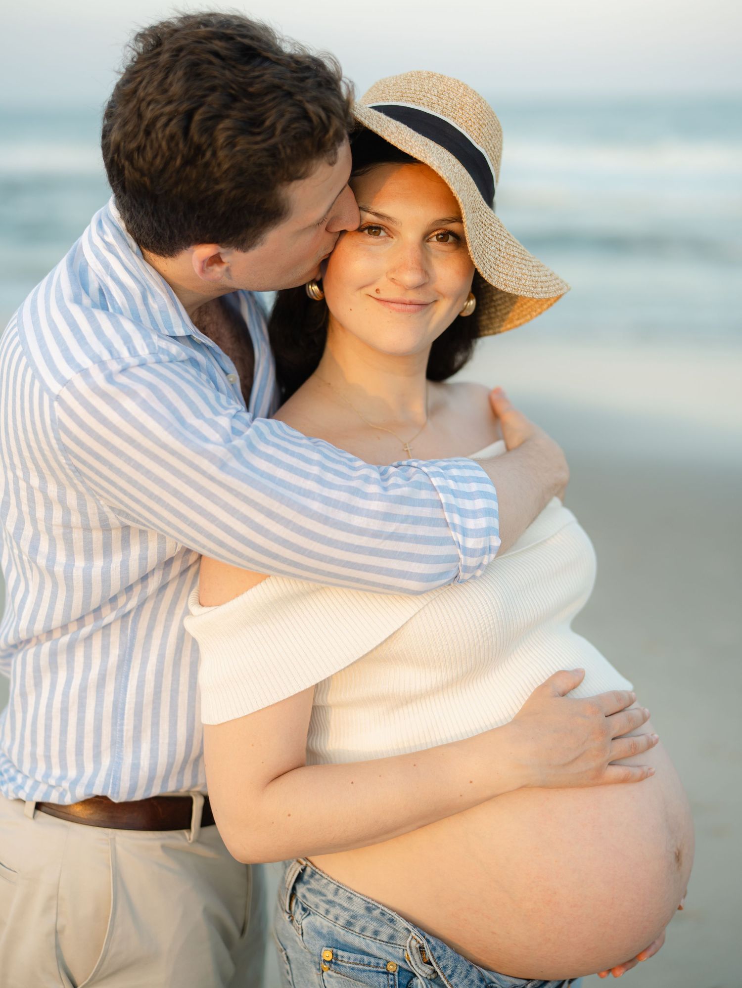 Pregnant woman with dark brown hair smiles as her husband kisses her.