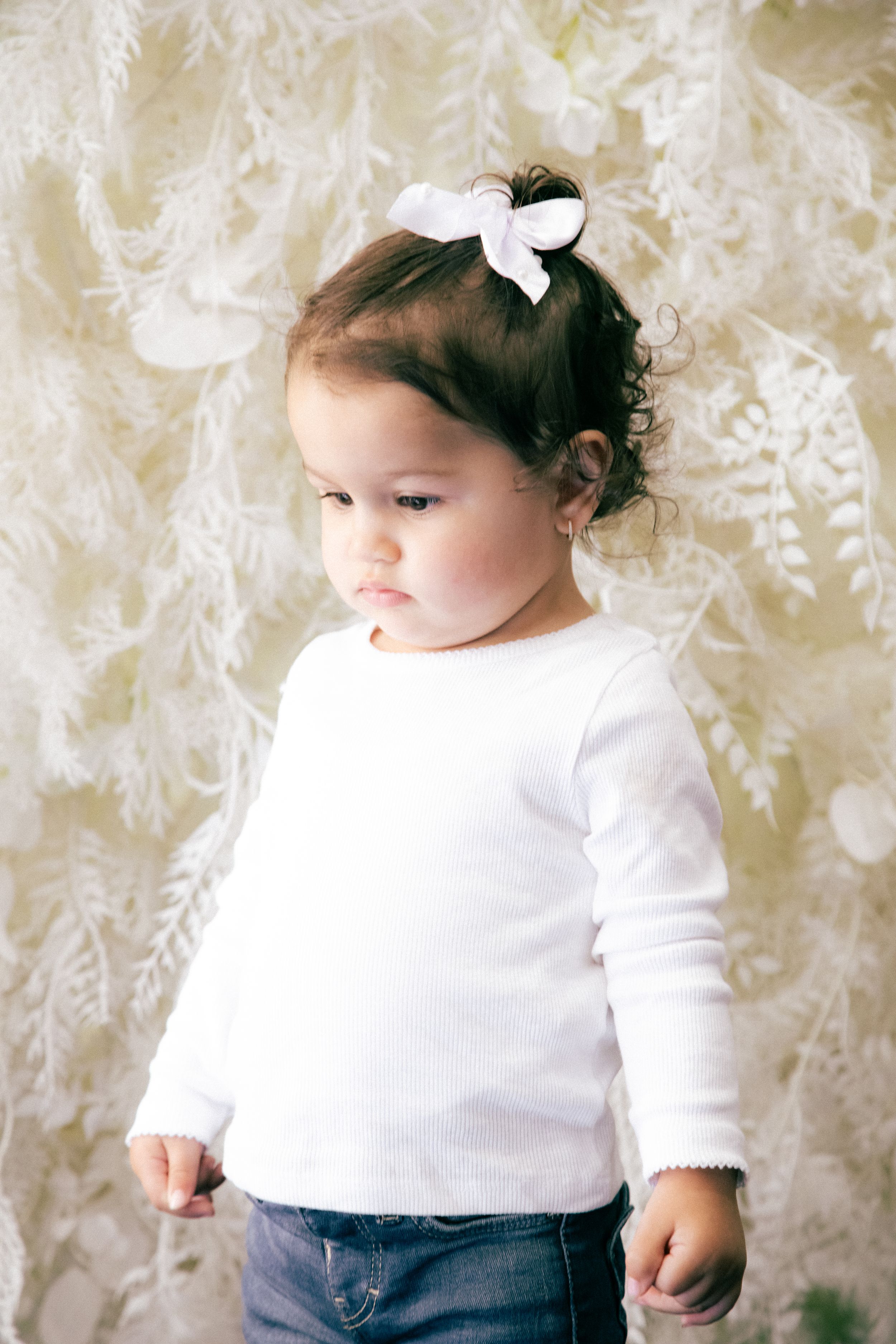 A toddler girl in a white long-sleeve shirt and jeans with a bow in her dark curly hair stands before a floral backdrop.