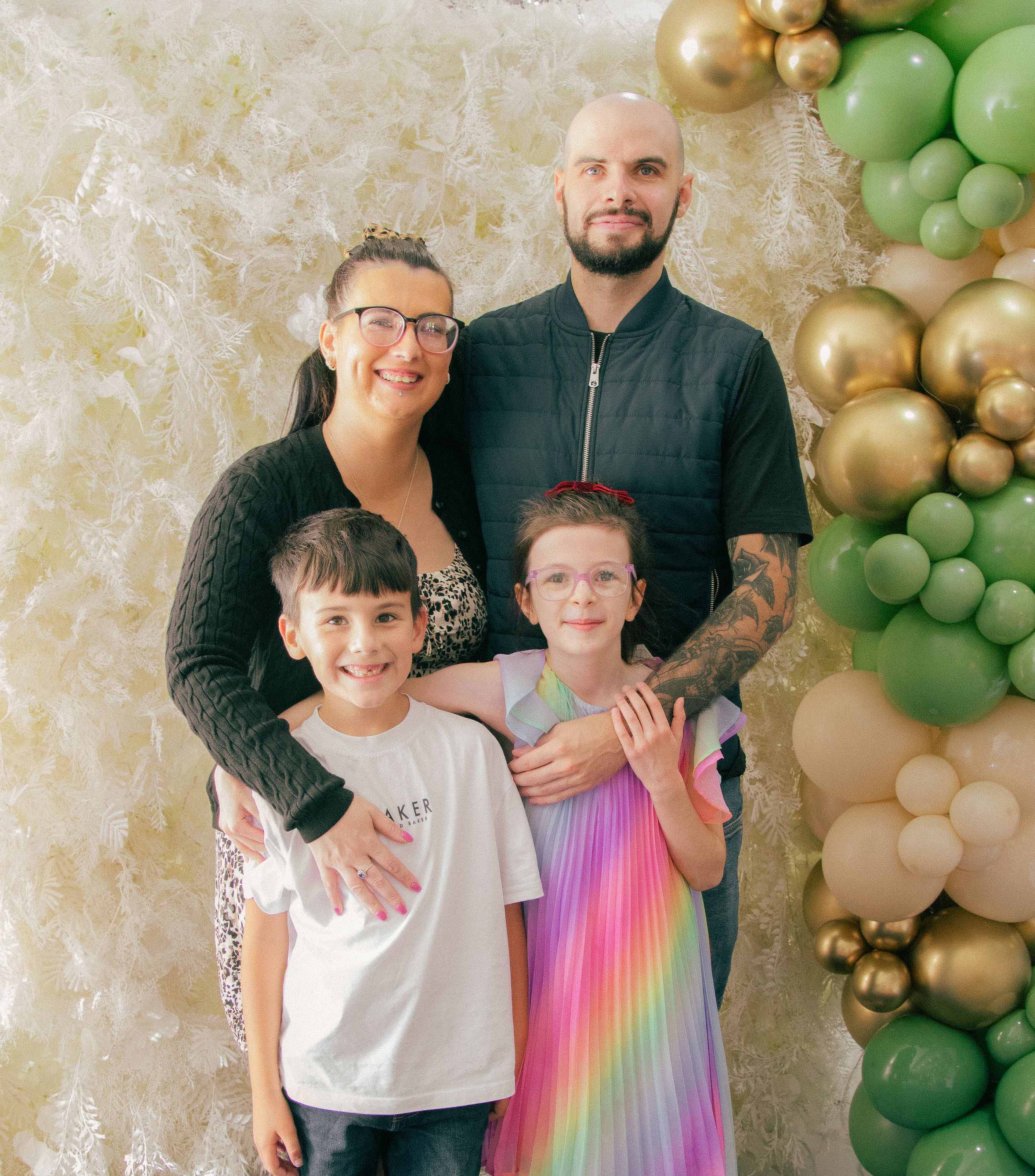 Family of four posing together in front of a balloon garland backdrop with green and gold balloons.