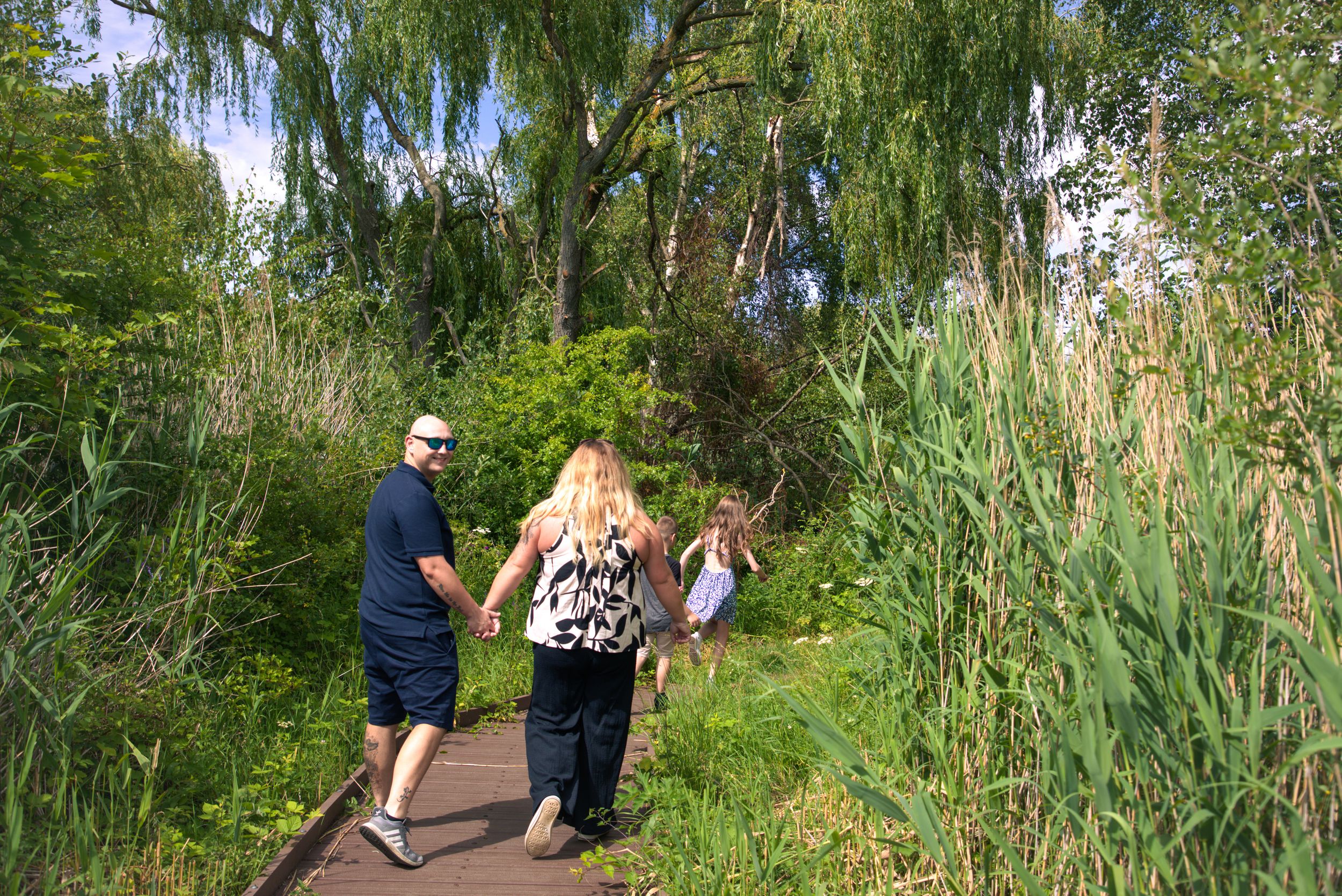 A family walks hand in hand along a wooden boardwalk path surrounded by lush green reeds and willow trees.