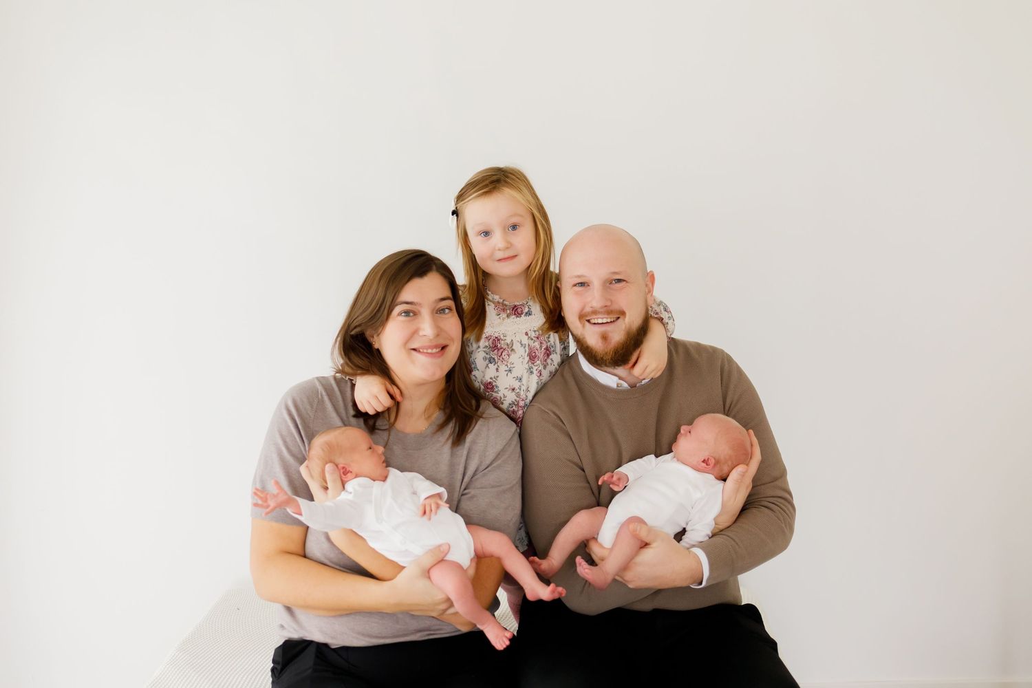 Happy family of four posing with newborn twins, smiling parents and young daughter in bright white studio.