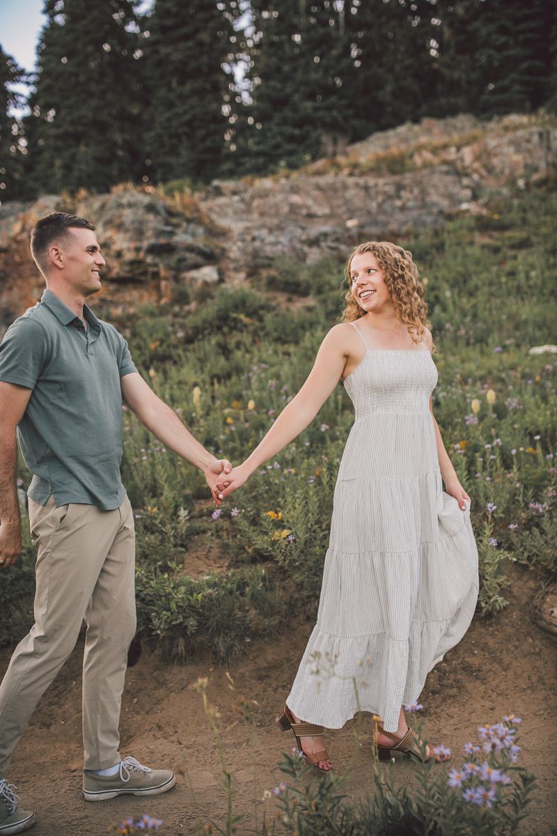 Couple holding hands on a mountain trail surrounded by wildflowers, woman in white dress smiling.