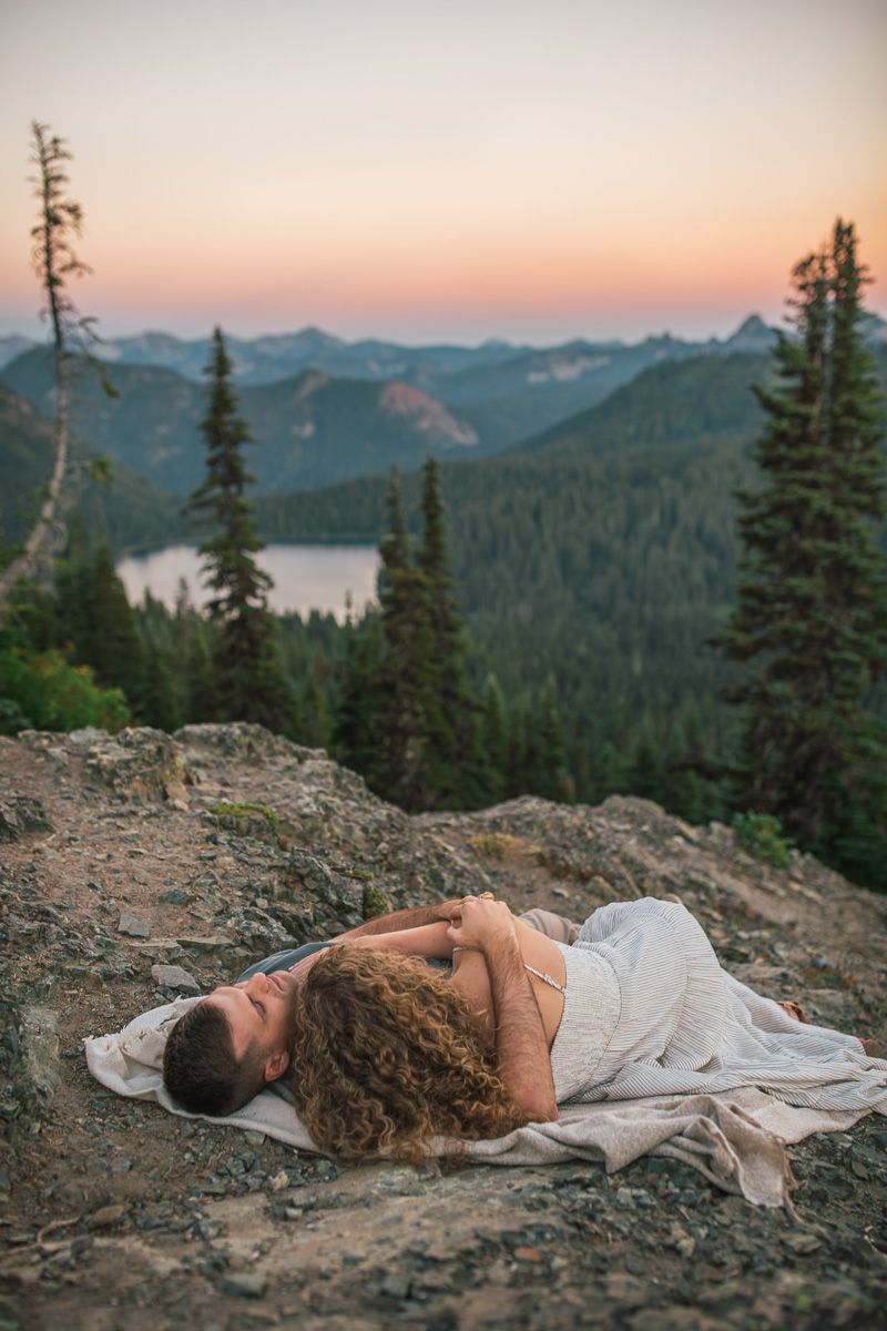 Couple lying together on rocky mountain overlook at sunset with lake and forest valley below.