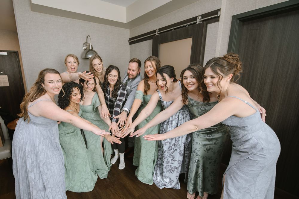 Bridesmaids in sage green dresses joining hands in a circle with the groom during wedding preparations indoors.