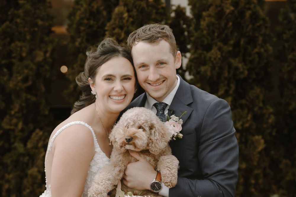 Smiling bride and groom holding a fluffy golden doodle dog on their wedding day outdoors.