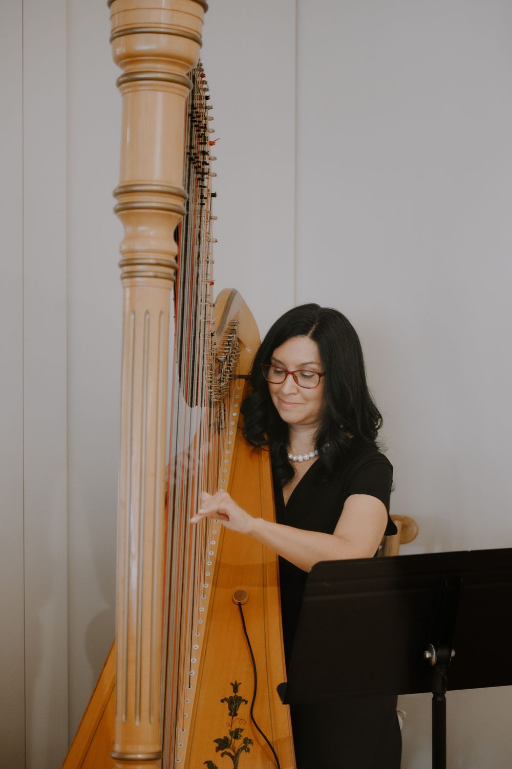 Woman in black dress playing a large wooden harp, wearing glasses and pearl necklace, standing by a music stand.