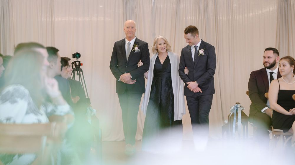 Groom escorted down the aisle by parents during indoor wedding ceremony with seated guests.