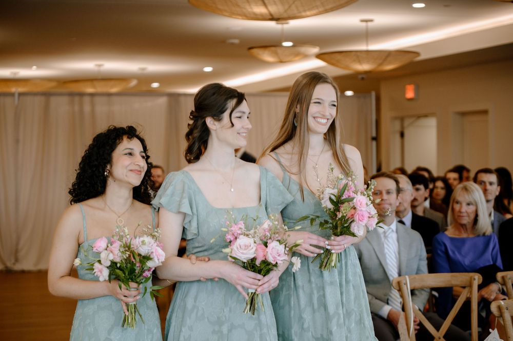 Three bridesmaids in sage green dresses carrying pink bouquets walk down the aisle at an elegant indoor wedding ceremony.
