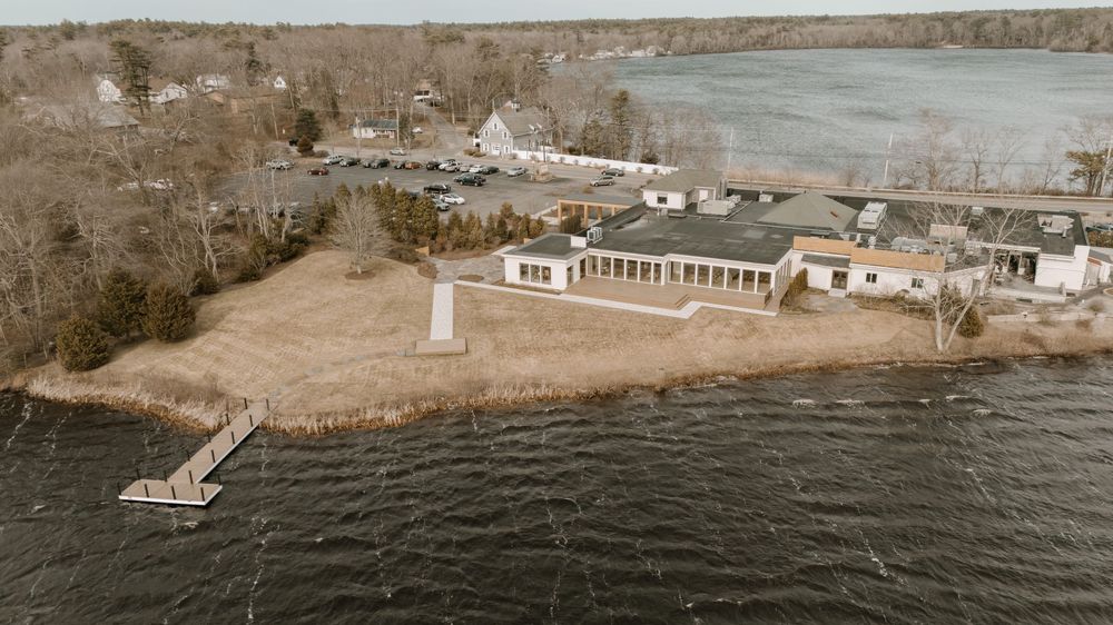 Aerial view of a waterfront wedding venue with a dock, monument, and reception hall beside a lake.