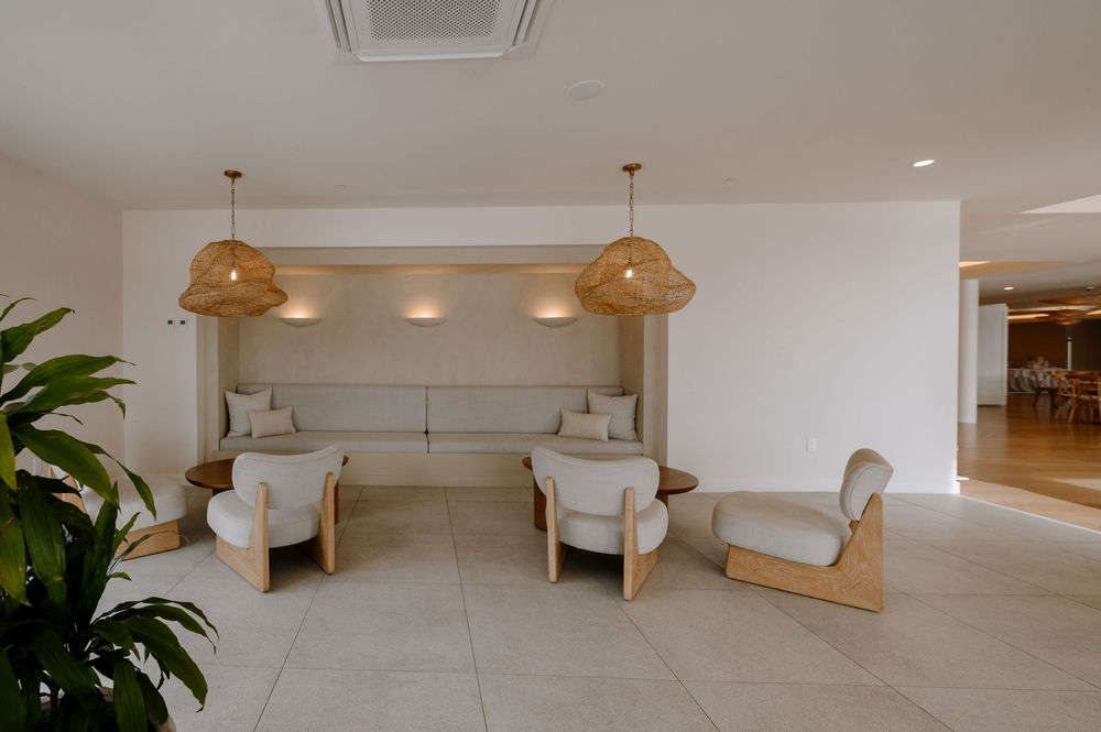 Modern hotel lounge with wicker pendant lights, white armchairs, and built-in sofa seating on beige tile floors.