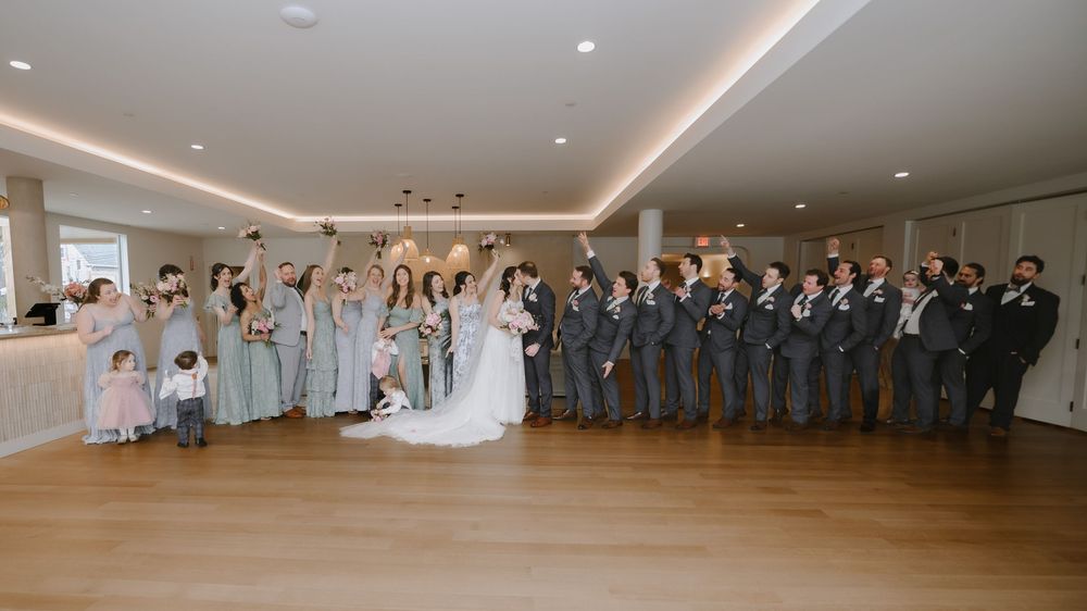 Large wedding party posing together in elegant venue with wood floors and neutral decor.