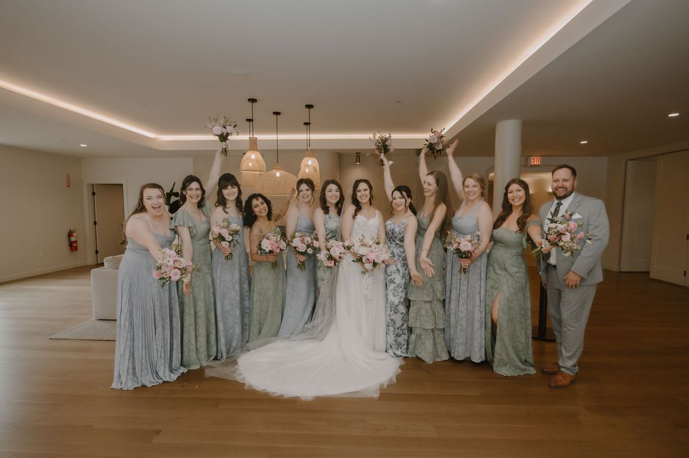 Bride in white gown poses with bridesmaids in sage green dresses and groomsman in gray suit indoors.