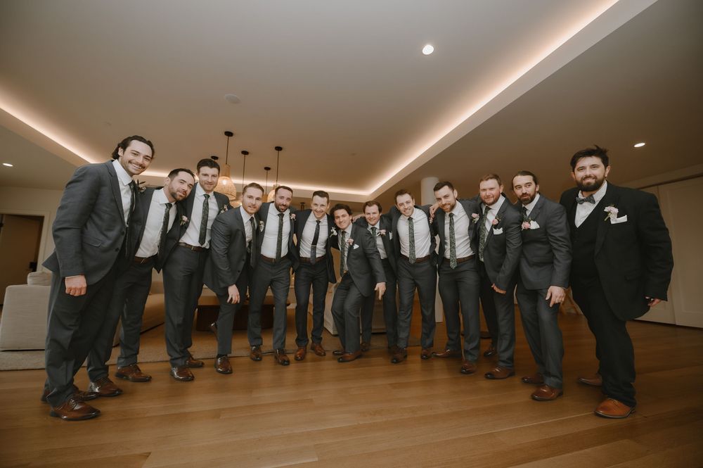 Groom poses with large groomsmen party in matching dark suits and ties in elegant venue.