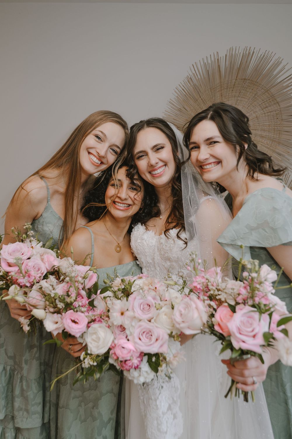 Bride in lace gown with three bridesmaids in sage green dresses holding pink rose bouquets.