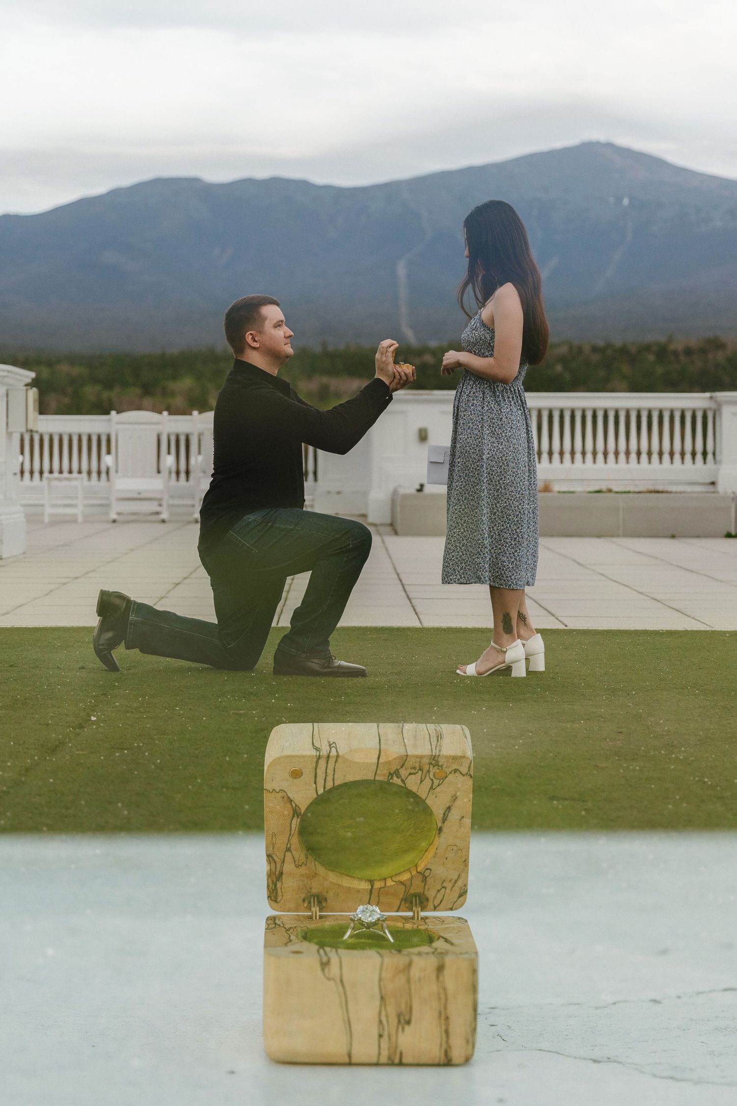 Man on one knee proposing to woman on rooftop terrace with mountain views and ring box in foreground.