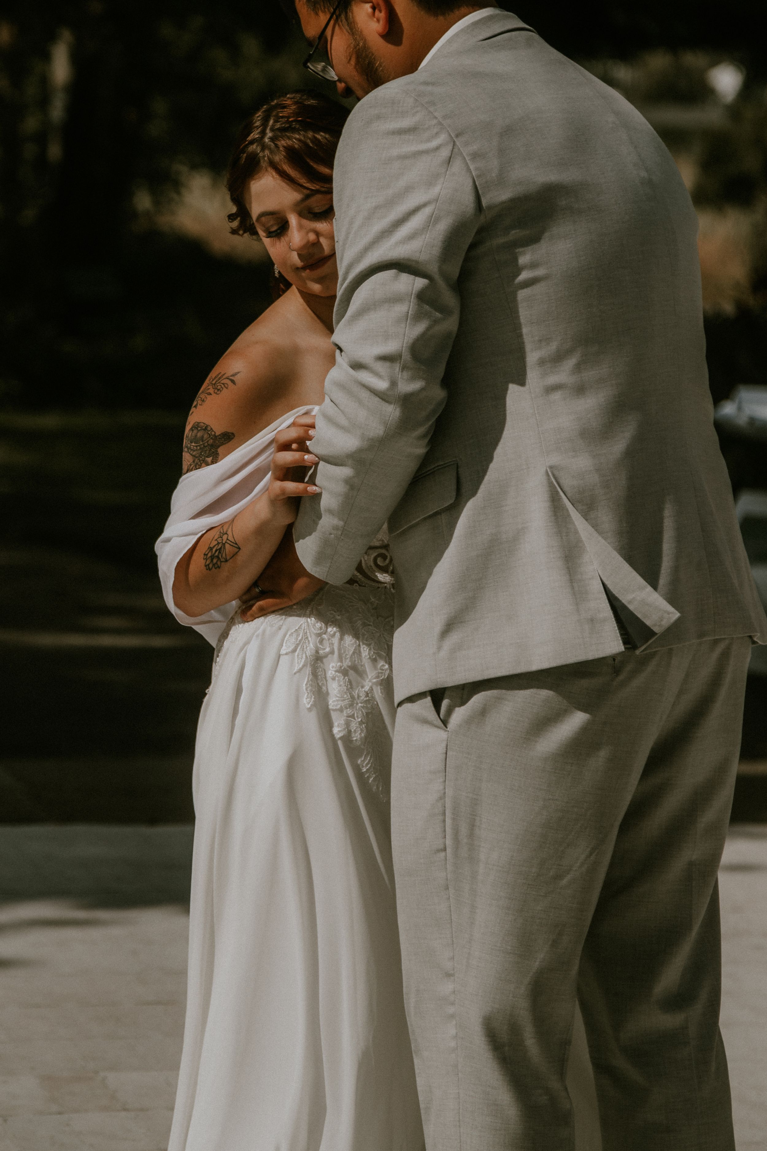 A bride and groom share an intimate moment, she in a white dress with tattoos, he in a light gray suit.