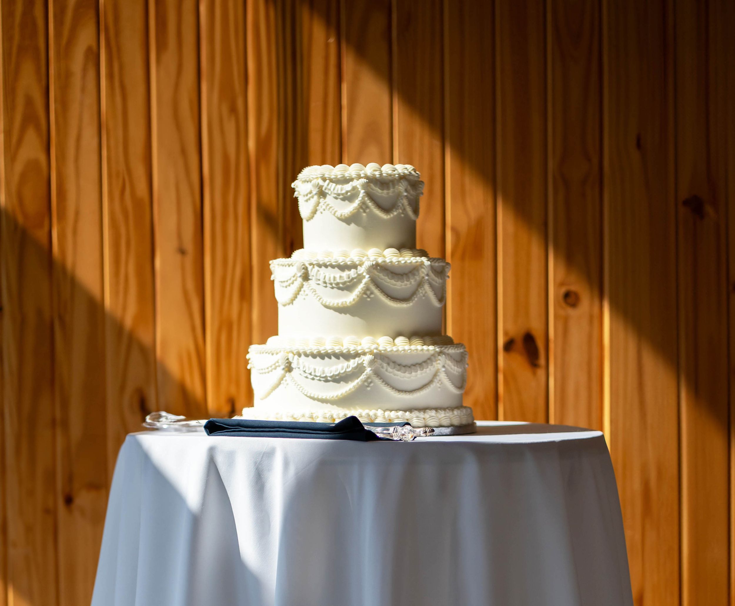 Elegant three-tier white wedding cake with decorative frosting on a draped table against a wood panel wall.